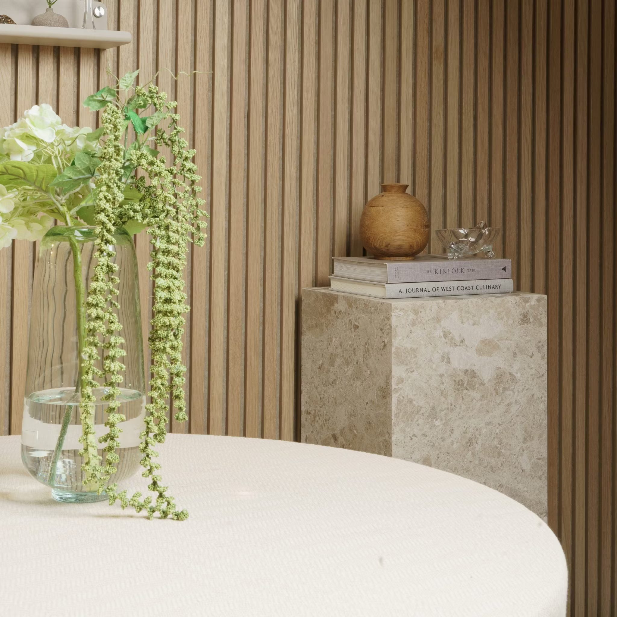 Eine Vase mit Blumen steht auf einem runden Tisch; Bücher und Töpferwaren ruhen auf einem Marmorsockel vor einer Wand aus Akupanel-Akustikholz.